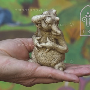 Pode incluir: Uma estatueta de gárgula bege com uma expressão caprichosa, segurada em uma mão. A gárgula tem uma superfície texturizada e está sentada. A imagem inclui o texto "TIBO GARGOYLES" e um logotipo com a palavra "TIBO".