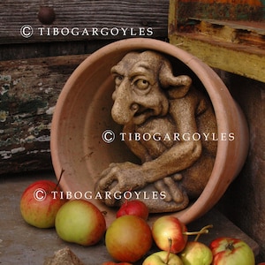 Puede incluir: Una maceta de color terracota con una detallada escultura de gárgola en su interior. Varias manzanas rojas y verdes están esparcidas en la base de la maceta. El texto "© TIBOGARGOYLES" es visible.