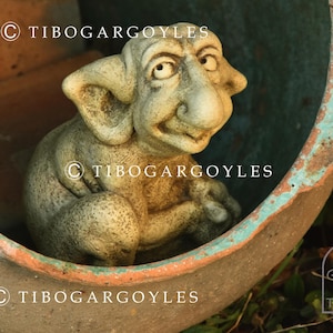 Op de afbeelding: Een verweerde stenen waterspuwer sculptuur met grote oren, een prominente neus en een lichte glimlach. De waterspuwer zit in een grote, verouderde terracotta pot met een turquoise rand. De tekst "© TIBOGARGOYLES" is inbegrepen.