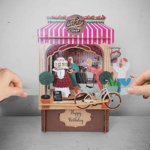 May include: A 3D paper cake shop scene with a pink and white striped awning, featuring figures holding cakes and a bicycle. The sign reads "Cake Shop" and "Happy Birthday".