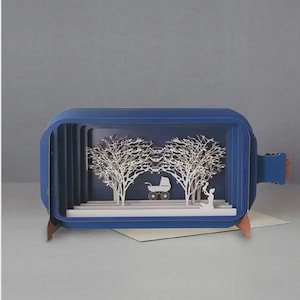 May include: A blue paper art card in the shape of a bottle, featuring a layered scene of white trees, a baby carriage, and a mother figure. The card is set against a gray background.