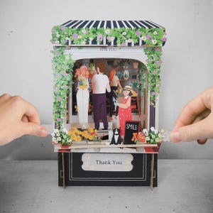 May include: A paper diorama of a flower shop with the text "FOR YOU" and "SMILE". The shop features illustrations of people, flowers, and a dog. The base of the shop has the text "Thank You".