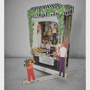 May include: A pop-up paper flower shop with a striped awning, floral accents, and the words "FOR YOU" and "SMILE." The shop features paper figures of people and flowers, with the text "Thank You" on the counter.
