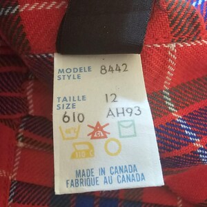May include: Close-up of a red, black, white, and blue plaid fabric with a tag attached. The tag reads "MODELE 8442 STYLE TAILLE 12 SIZE 610 AH93" and includes care symbols. The text "MADE IN CANADA FABRIQUE AU CANADA" is also visible.