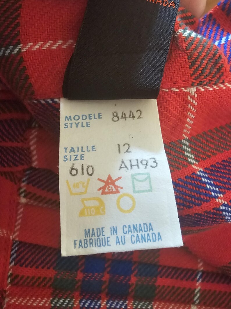 May include: Close-up of a red, black, white, and blue plaid fabric with a tag attached. The tag reads "MODELE 8442 STYLE TAILLE 12 SIZE 610 AH93" and includes care symbols. The text "MADE IN CANADA FABRIQUE AU CANADA" is also visible.