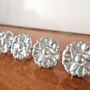 Puede incluir: Cuatro tiradores de armario plateados y ornamentados con un diseño floral. Los tiradores están dispuestos en fila, con dos tiradores en primer plano y dos en segundo plano. Los tiradores son de metal y tienen un acabado ligeramente envejecido.