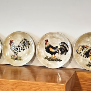 Peut inclure: Quatre assiettes décoratives avec des motifs de coqs peints à la main. Chaque assiette présente un coq différent avec des couleurs vives, dont le rouge, le noir, le jaune et le blanc, sur un fond neutre. Les assiettes sont exposées sur une surface en bois.