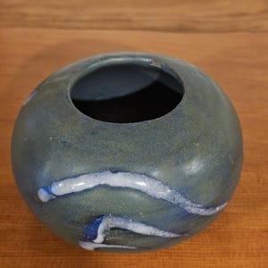 Peut inclure: Un petit vase rond en céramique dans les tons de bleu et de vert. Le vase a une large ouverture et est décoré de lignes ondulées blanches et bleues. La poterie faite à la main a une finition mate et convient pour exposer de petites fleurs ou comme élément décoratif.