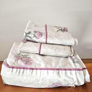 May include: Folded bed linen featuring a floral design. The sheets are a pale beige with a pattern of pink flowers and green foliage. A dark pink border edges the sheets, creating a contrasting detail.