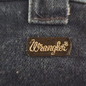 May include: Close-up of dark blue denim fabric with a rectangular black label. The label features the brand name "Wrangler" in white cursive script. The denim has a textured appearance, and a portion of a seam is visible.