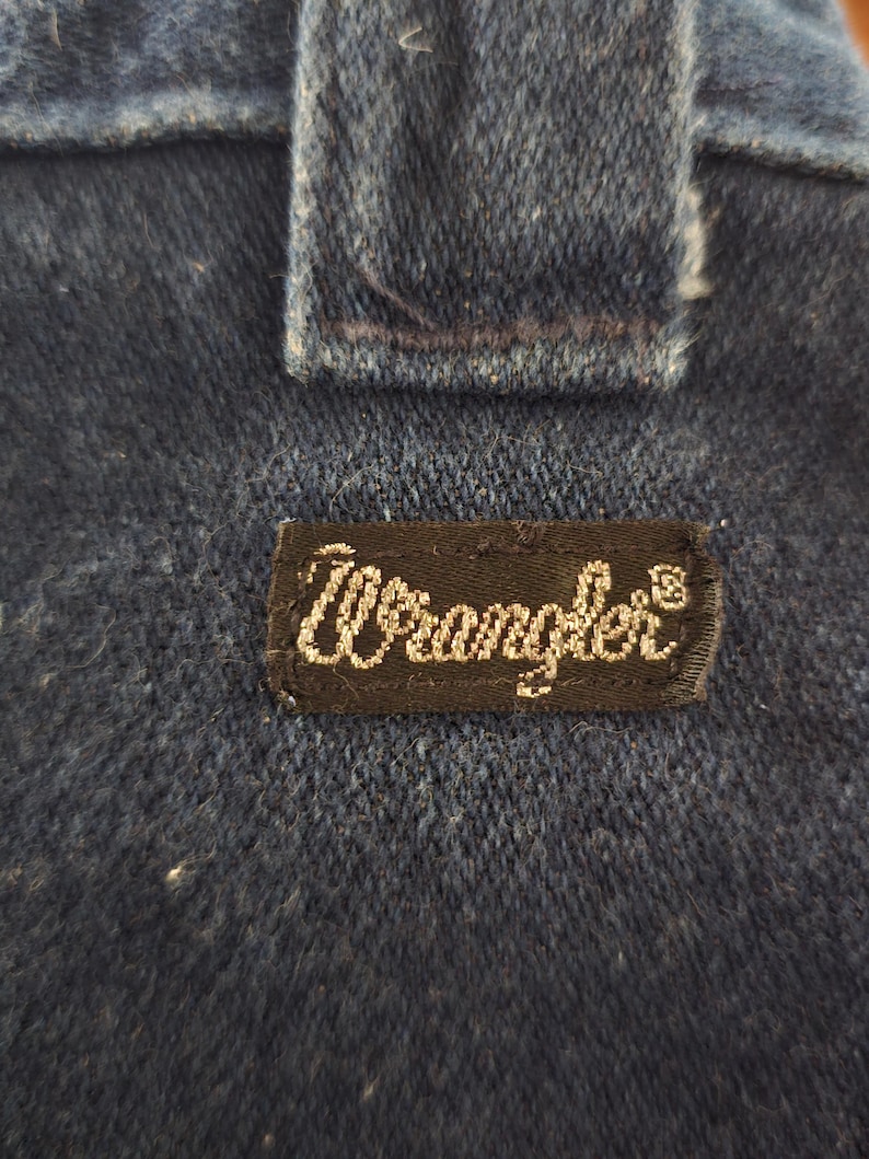 May include: Close-up of dark blue denim fabric with a rectangular black label. The label features the brand name "Wrangler" in white cursive script. The denim has a textured appearance, and a portion of a seam is visible.