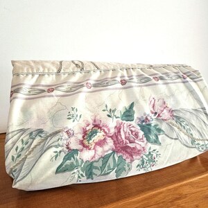 Könnte beinhalten: Eine Clutch mit Blumenmuster auf cremefarbenem Hintergrund. Sie zeigt große rosa Rosen, grüne Blätter und einen dekorativen Rand mit Seilmotiv. Der Stoff hat eine Vintage-Ästhetik.