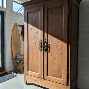 以下が含まれることがあります： 2つのドアと装飾的な金属製のハンドルが付いた、大きなアンティークの木製キャビネット。キャビネットは温かみのある茶色で、コンクリートの床に立っています。キャビネットは、サーフボードとタオルが見える明るい部屋にあります。