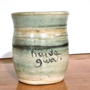May include: Handmade ceramic mug with a cylindrical form. The mug displays a mix of green, beige, and brown tones, with visible brushstrokes. The words "harda gwa" are inscribed on the side in black.