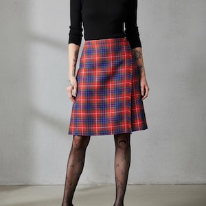 May include: A red, blue, and white plaid skirt with a wrap design. The skirt is paired with a black turtleneck and patterned tights. The outfit is completed with black high heels.