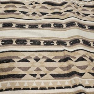 May include: A woven textile with a geometric pattern in shades of cream, tan, brown, and black. The design features horizontal stripes, triangles, and diamond shapes, creating a textured, Southwestern-inspired look. The fabric appears soft and possibly wool or a wool blend.