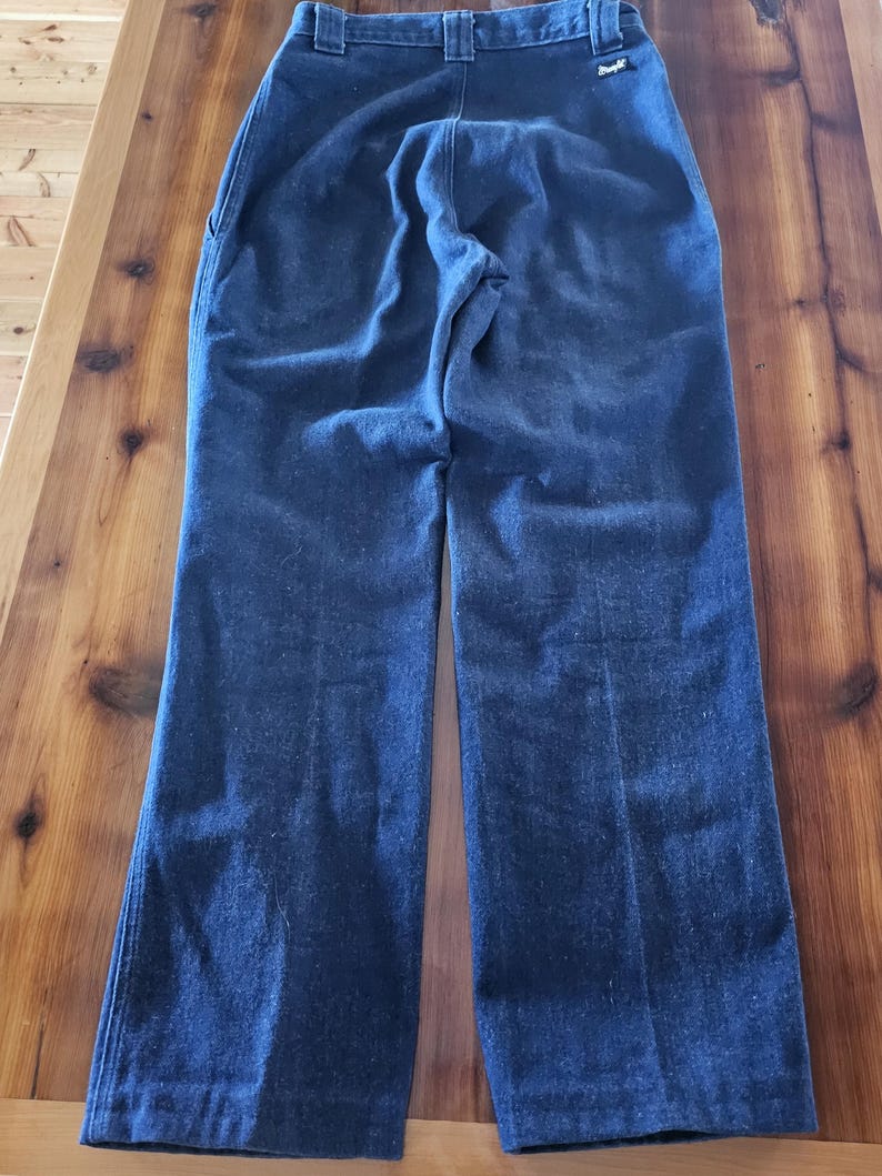 May include: Dark blue denim pants with a high waist and a straight leg. The pants have a classic five-pocket design and a Wrangler logo on the back pocket. The pants are made of a sturdy denim material.