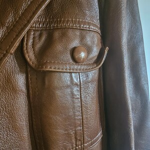 May include: Close-up of a brown leather jacket. The jacket has a pocket with a button and detailed stitching. The leather has a textured appearance, suggesting a vintage or classic style. The jacket is brown.