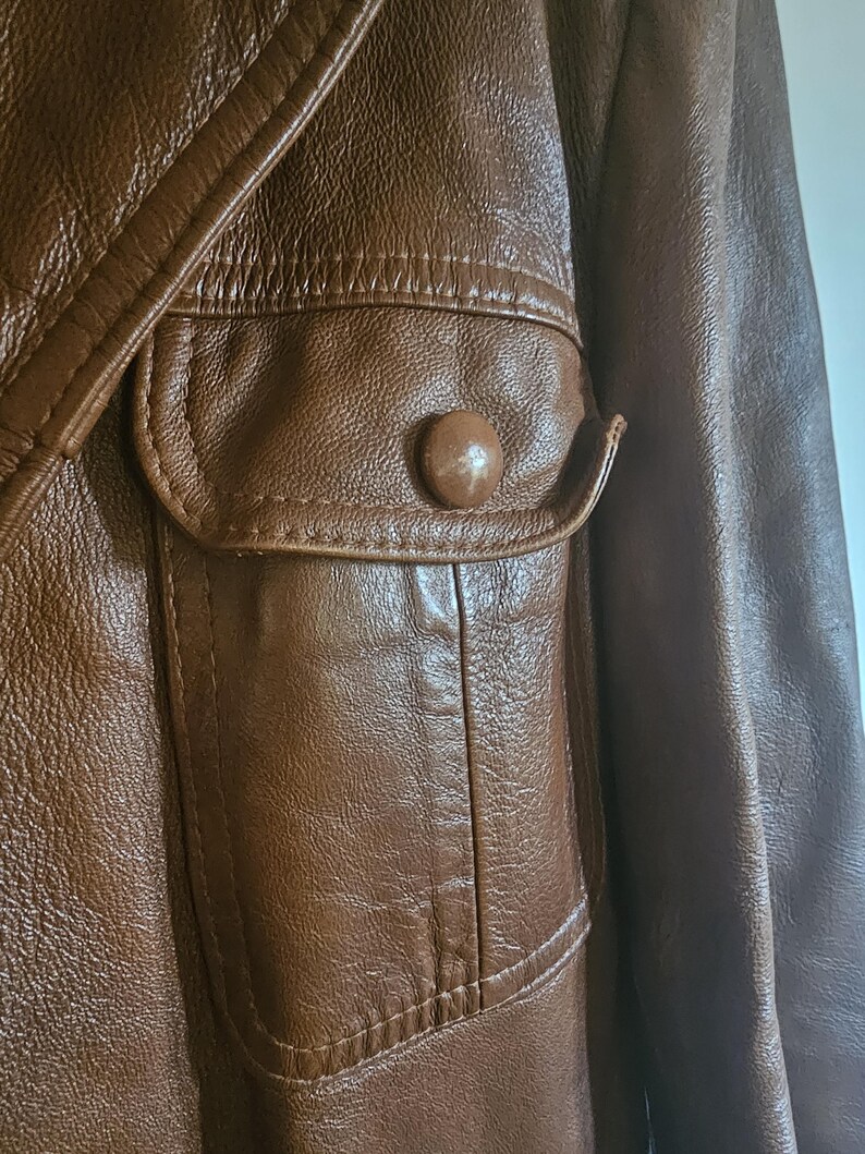 May include: Close-up of a brown leather jacket. The jacket has a pocket with a button and detailed stitching. The leather has a textured appearance, suggesting a vintage or classic style. The jacket is brown.