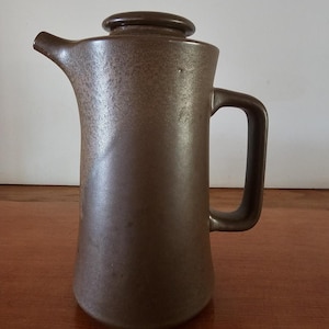 May include: A brown ceramic coffee pot with a lid and handle. The pot has a spout for pouring and a cylindrical body that tapers slightly towards the base. The surface has a speckled texture.