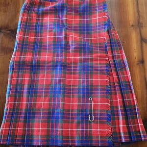 May include: A red, blue, and white plaid kilt with a fringed edge and a safety pin closure. The kilt is made of a woven fabric and has a pleated design. The kilt is displayed on a wooden surface.