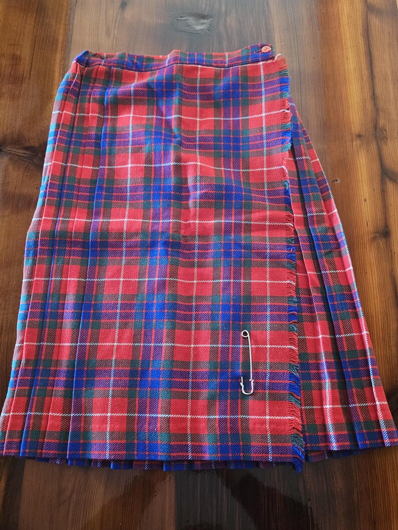 May include: A red, blue, and white plaid kilt with a fringed edge and a safety pin closure. The kilt is made of a woven fabric and has a pleated design. The kilt is displayed on a wooden surface.