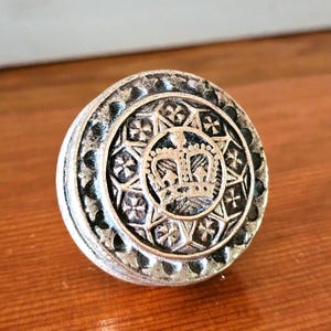 May include: A silver-toned, circular button with a detailed crown design at the center. The button features an outer ring with a repeating pattern of crosses and stars. The button is likely made of metal and has a slightly aged appearance.