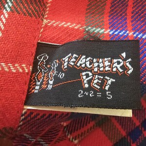 May include: Close-up of a red plaid fabric with a black label. The label reads "Teacher's Pet" in white lettering with an illustration and the equation "2+2=5". The plaid pattern includes red, blue, green, and white lines.