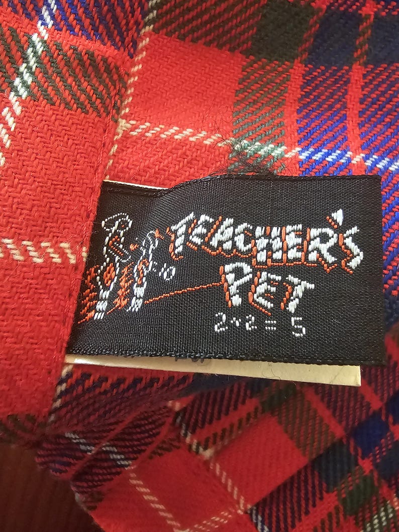 May include: Close-up of a red plaid fabric with a black label. The label reads "Teacher's Pet" in white lettering with an illustration and the equation "2+2=5". The plaid pattern includes red, blue, green, and white lines.