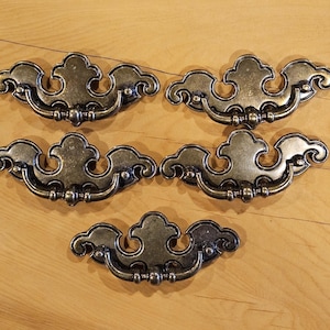 May include: Five antique-style drawer pulls with a cloud-like design. The pulls are made of a dark metal with a slightly tarnished finish. Each pull has a curved handle and decorative detailing.