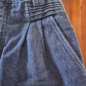 May include: Close-up of a pair of blue denim shorts. The shorts feature a button closure, belt loops, and pleats at the waist. The fabric has a textured appearance, and the shorts appear to be vintage.