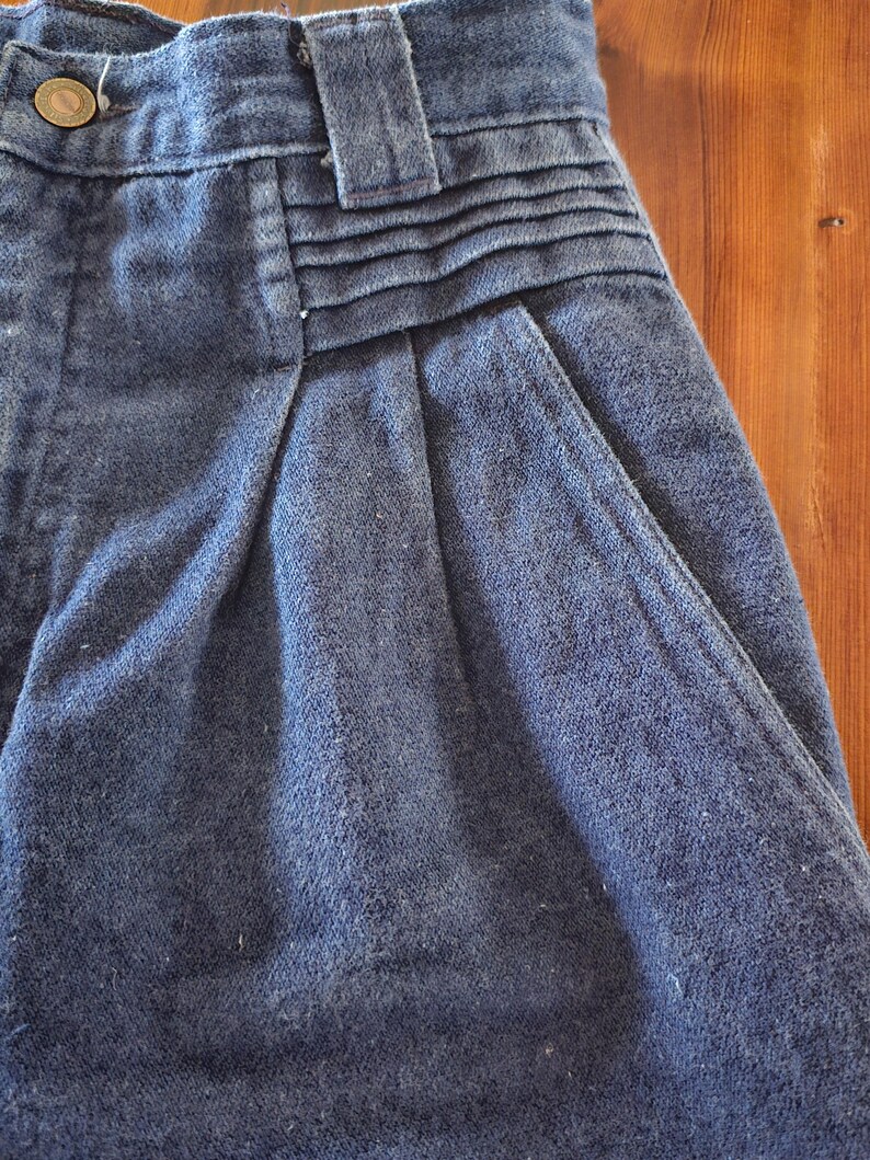 May include: Close-up of a pair of blue denim shorts. The shorts feature a button closure, belt loops, and pleats at the waist. The fabric has a textured appearance, and the shorts appear to be vintage.