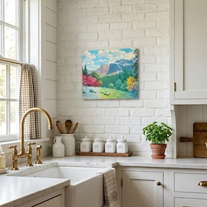May include: A framed landscape painting depicting a mountain range, green trees, and a field of pink flowers hangs on a white brick wall in a kitchen. The kitchen features a white sink, gold faucet, and white cabinets.