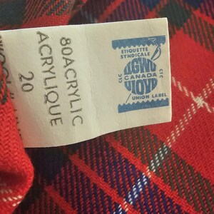 May include: Close-up of a red, blue, and white plaid fabric with a tag. The tag reads "80 Acrylic" and "Union Label" with a blue and white logo. The fabric has a woven texture.