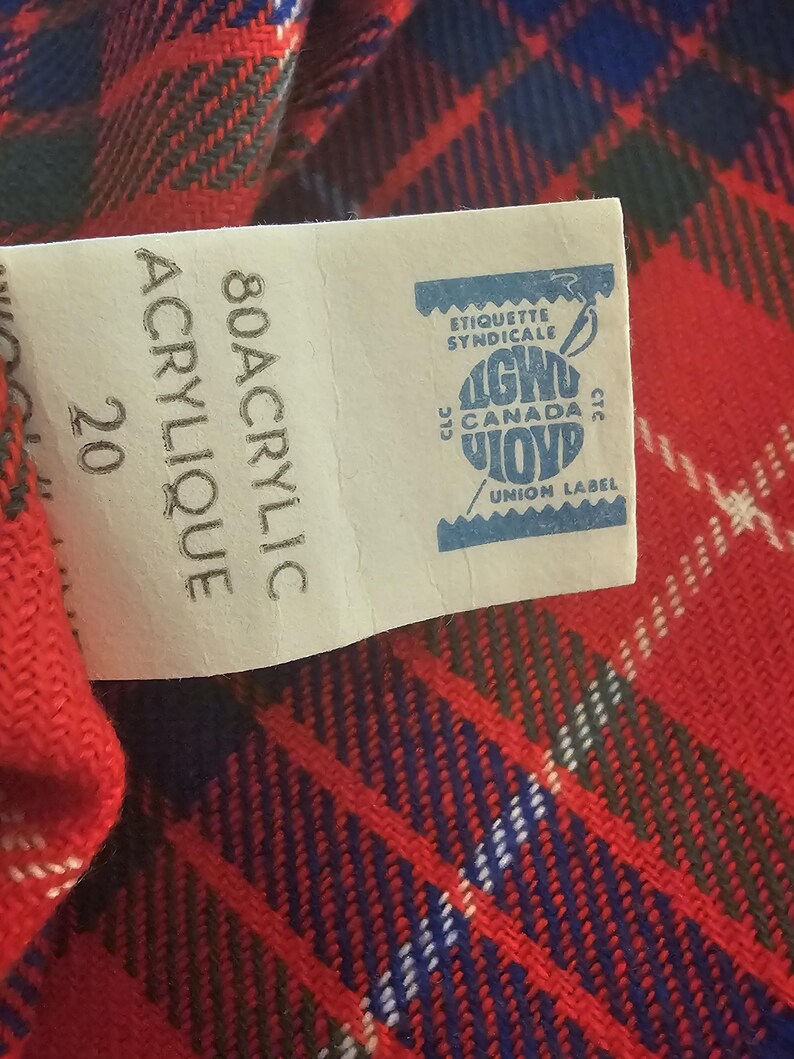 May include: Close-up of a red, blue, and white plaid fabric with a tag. The tag reads "80 Acrylic" and "Union Label" with a blue and white logo. The fabric has a woven texture.