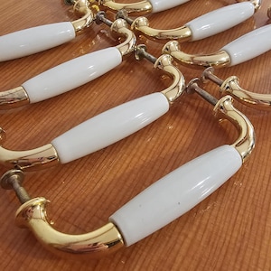May include: A selection of cabinet handles featuring a gold-coloured finish and white ceramic centre. The handles have a curved design, and each includes a screw for fitting. The hardware is arranged on a wooden surface.
