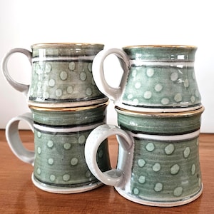 May include: Four stacked ceramic mugs with a green and white polka dot pattern. Each mug has a gray handle and a gold rim. The mugs are arranged on a wooden surface.