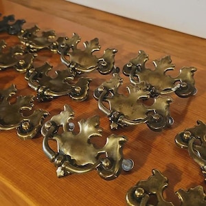 以下が含まれることがあります： アンティーク真鍮製の引き出し用取っ手のコレクション。葉のような装飾的なデザインが特徴です。取っ手は曲線的なハンドルと装飾的なバックプレートを備え、ヴィンテージ感を演出しています。木製の表面に配置されています。