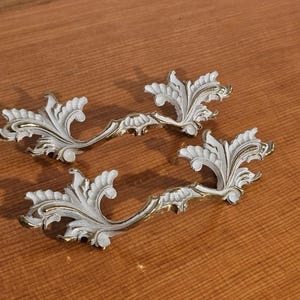 Könnte beinhalten: Zwei weiß-goldene dekorative Schubladengriffe mit floralem Design. Die Griffe haben eine gebogene Form und sind aus Metall gefertigt. Die weißen Bereiche haben eine strukturierte Optik, und die goldenen Akzente verleihen einen Hauch von Eleganz.