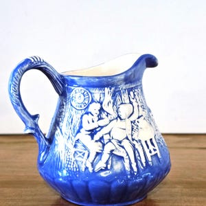 May include: A blue and white ceramic creamer pitcher with a raised design of people and a clock. The pitcher has a handle and a rounded body.