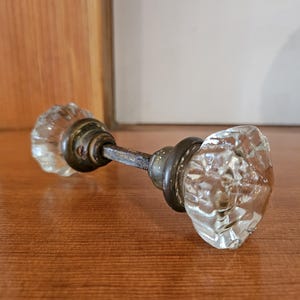 May include: A door knob set featuring two clear, faceted glass knobs connected by a central metal spindle. Each glass knob is attached to a circular, dark-toned metal base plate, showing signs of age and wear on the metal components.