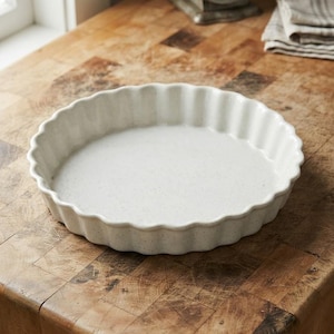 Peut inclure: Un moule à tarte rond en céramique blanc cassé avec une finition mouchetée et un bord cannelé. L'ustensile présente une base plate peu profonde et des parois verticales ondulées, offrant une apparence neutre et épurée.