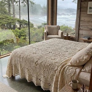 May include: A bedroom with a large window offering a coastal view. The bed is adorned with a cream-coloured, intricately crocheted blanket. A matching pillow rests on the bed. A wooden bedside table holds a mug and a book.