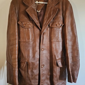 May include: A brown leather jacket with four front pockets and a notched collar. The jacket has a three-button closure and a tailored fit. The jacket is hanging on a white hanger.
