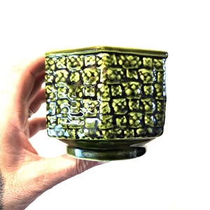 May include: A square, green ceramic planter with a textured, grid-like pattern. The planter has a small, circular base and a slightly flared rim. The dark green glaze gives it a glossy finish. A hand is holding the planter.