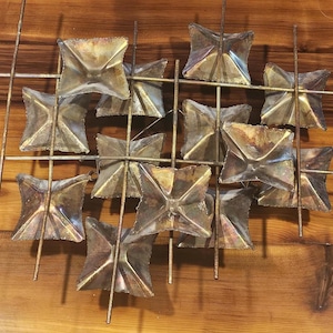 May include: A vintage metal wall sculpture featuring a grid of folded, square shapes in shades of silver and bronze. The squares are arranged in a grid pattern, with thin metal rods connecting them. The sculpture is mounted on a wooden surface.