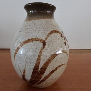 May include: A ceramic vase with a rounded body and a narrow neck. The base colour is a speckled off-white, featuring hand-painted brown brushstrokes that depict stylised grass or floral elements. The rim and neck are finished in a solid brown glaze.