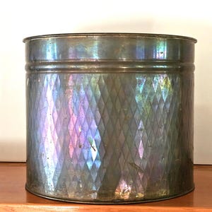 May include: A cylindrical metal container with a diamond pattern. The container has a metallic sheen with iridescent colours of blue, purple, and green. The container is on a wooden surface.