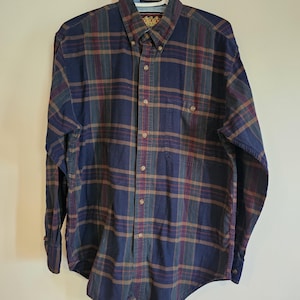 May include: A long-sleeved, navy blue, burgundy, and olive green plaid shirt. It has a button-down front, a chest pocket, and a classic collar. The shirt is displayed on a white hanger.