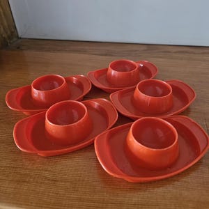 May include: Six red plastic egg cups, each with a circular base and a central cup to hold an egg. The egg cups are arranged on a wooden surface, showcasing their bright color and simple design. These are ideal for serving eggs.
